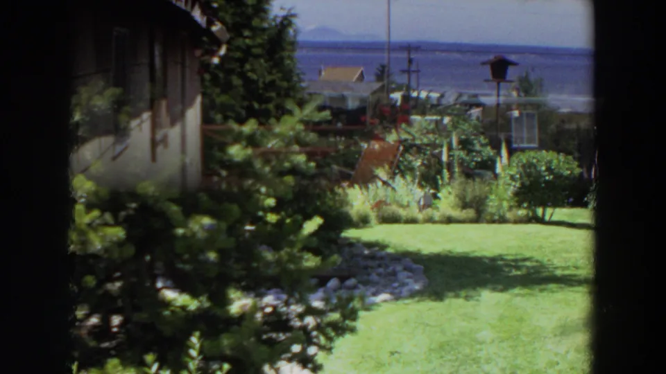 1964 Washington home-movie frame showing a lawn, shrubs, house wall, red flowers, and a partial deck view toward distant water.