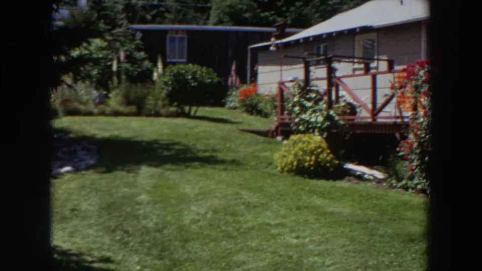1964 Washington garden scene with a sunlit lawn, flowering bushes, trees, and a modest house with wooden deck railings.