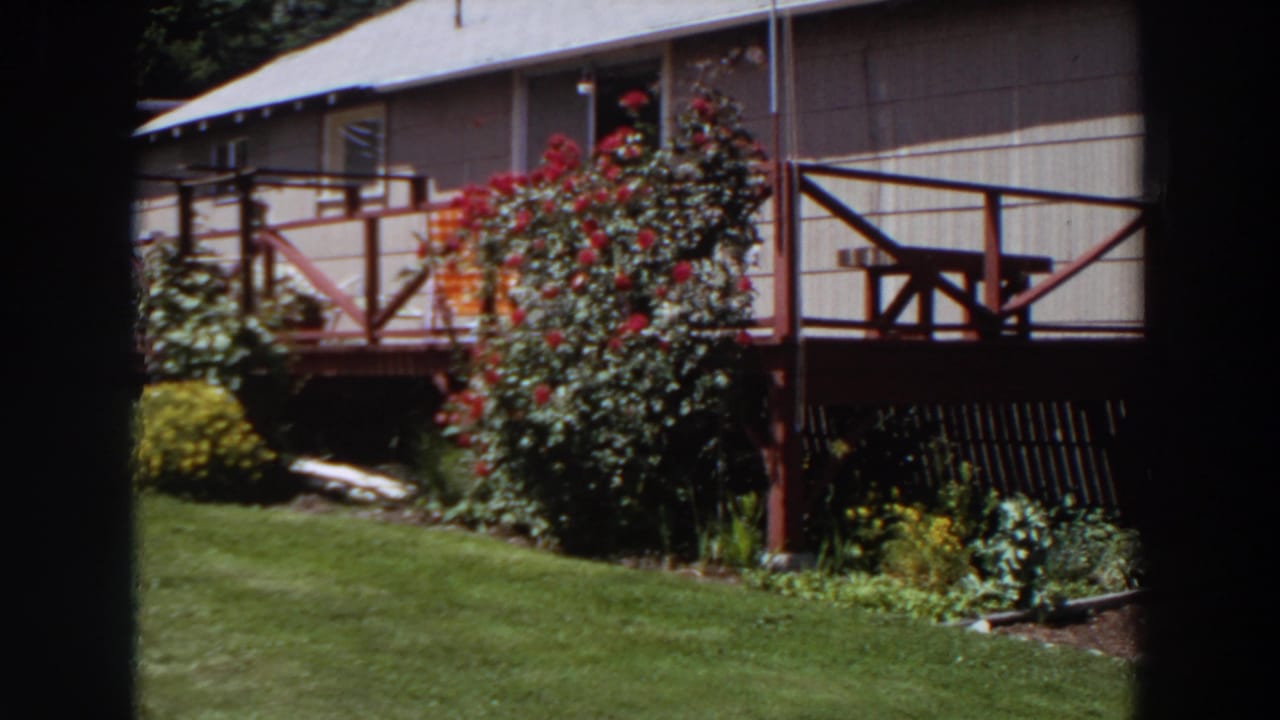 1964 Washington home-movie frame showing a house deck with cross railings, red flowers, shrubs, lawn, and garden planting.