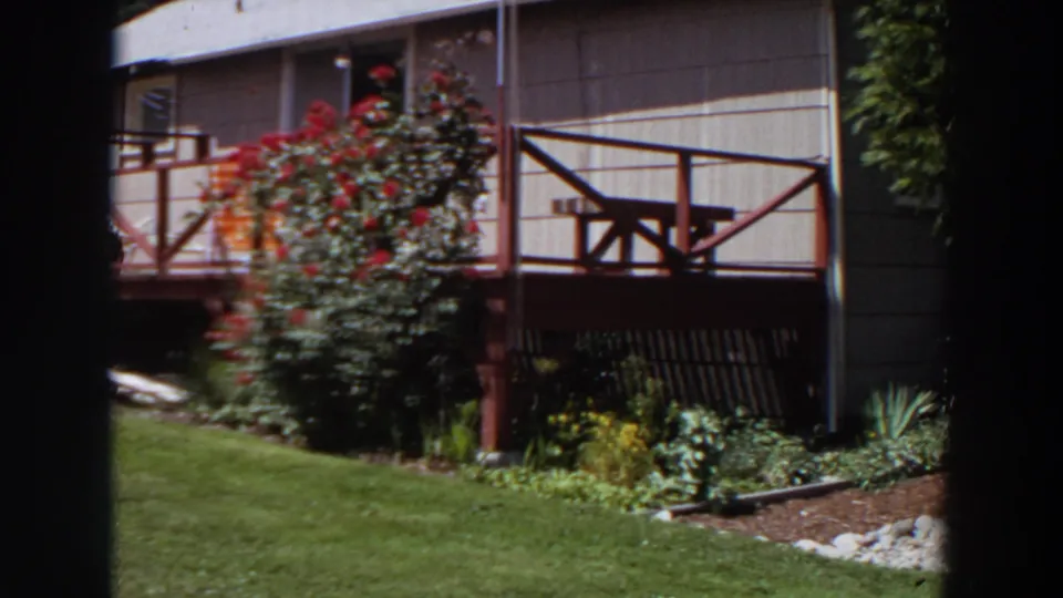 1964 Washington 8mm frame showing a pale rural house, red porch railing, picnic table, lawn, and flowering garden beds.