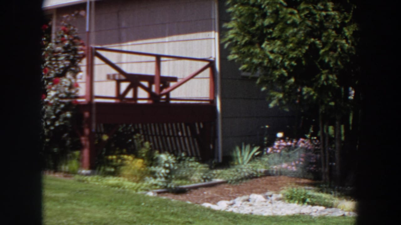 1964 Washington home-movie frame with a red porch, pale siding, landscaped beds, green lawn, and a leafy tree at the yard edge.