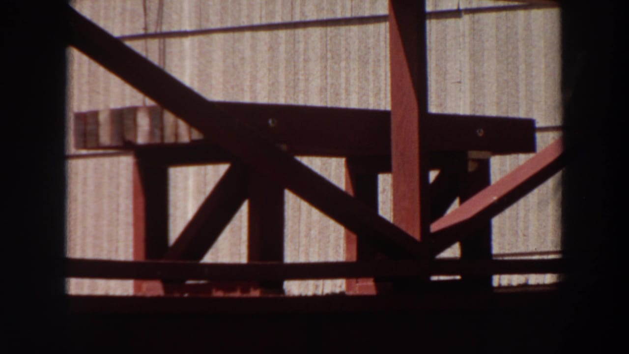 1964 Washington home-movie frame shifts to a nearby wood deck railing and vertical exterior siding viewed through a dark window frame.