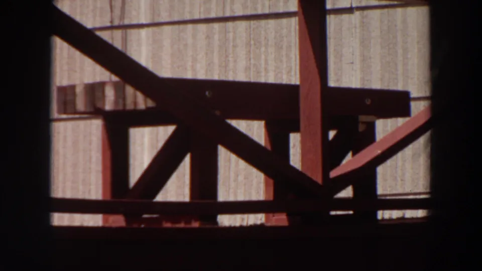 1964 Washington home-movie frame shifts to a nearby wood deck railing and vertical exterior siding viewed through a dark window frame.