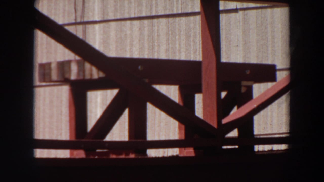 1964 Washington home-movie frame with red wooden rails, diagonal bracing, and a pale grooved wall behind them.