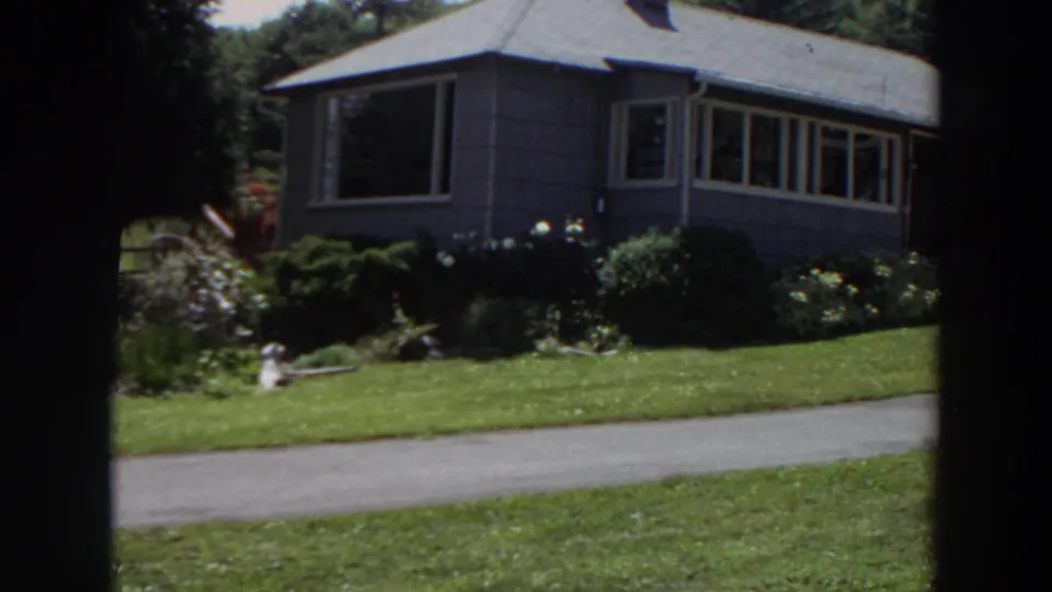 1964 Washington 8mm frame showing a grey-sided house, front lawn, driveway, shrubs, and white-flowered garden beds.