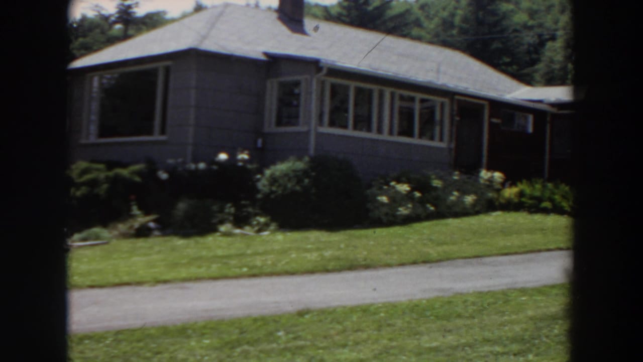 1964 Washington home-movie frame of a grey house exterior with a neat garden, green lawn, and driveway crossing the foreground.