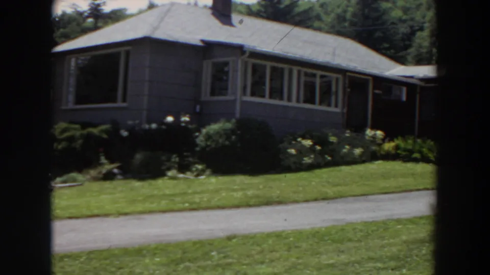 1964 Washington home-movie frame of a grey house exterior with a neat garden, green lawn, and driveway crossing the foreground.