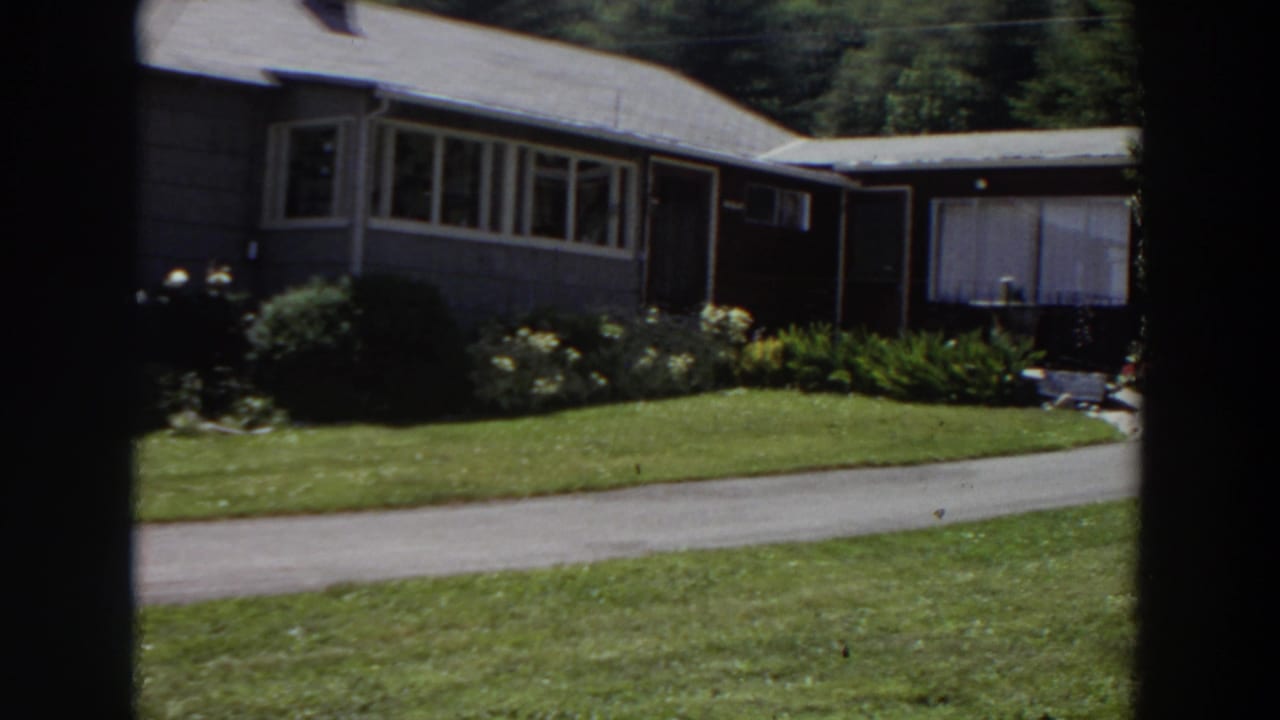 1964 Washington 8mm frame showing the pan reaching the house side, with long windows, flower beds, lawn, and driveway visible.