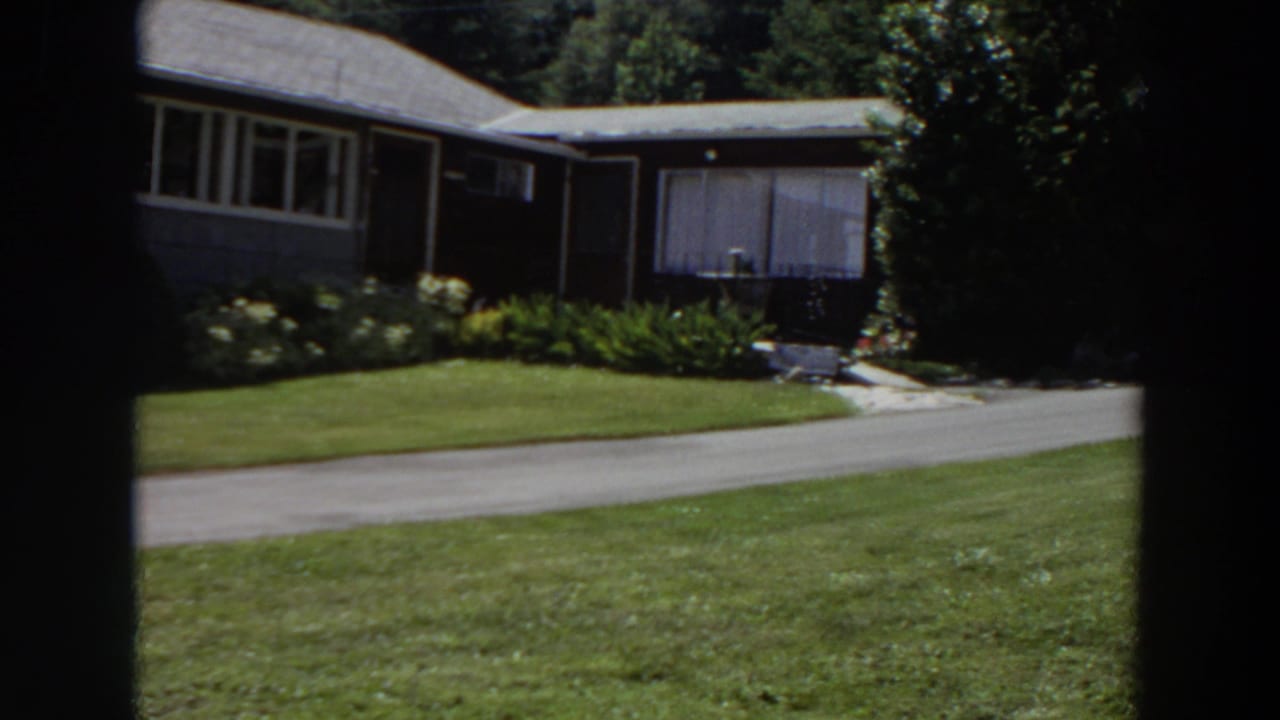1964 Washington home-movie frame of the house side, adjoining structure, driveway, clipped lawn, garden plants, and tree cover.