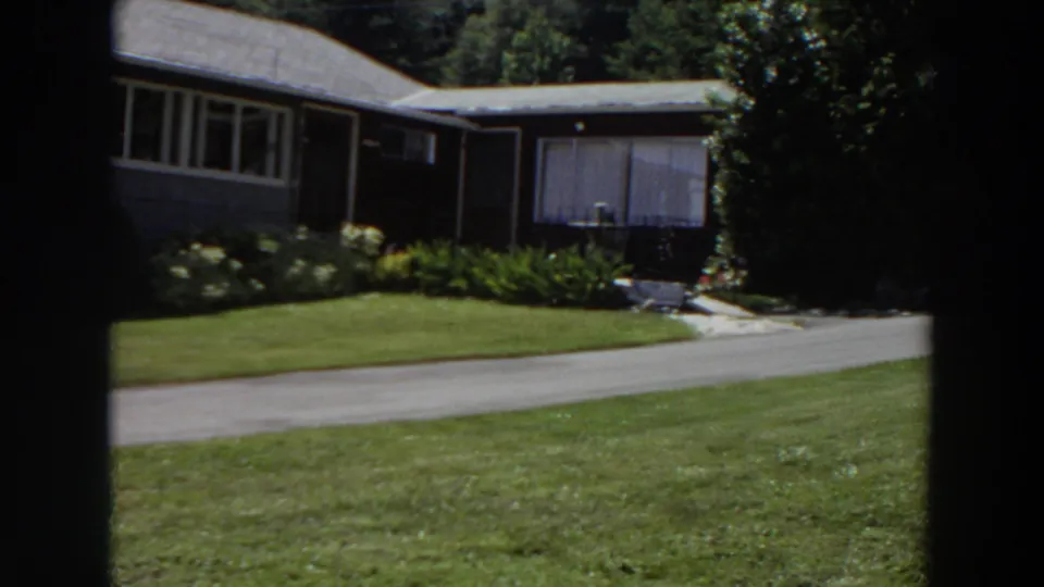 1964 Washington home-movie frame of the house side, adjoining structure, driveway, clipped lawn, garden plants, and tree cover.