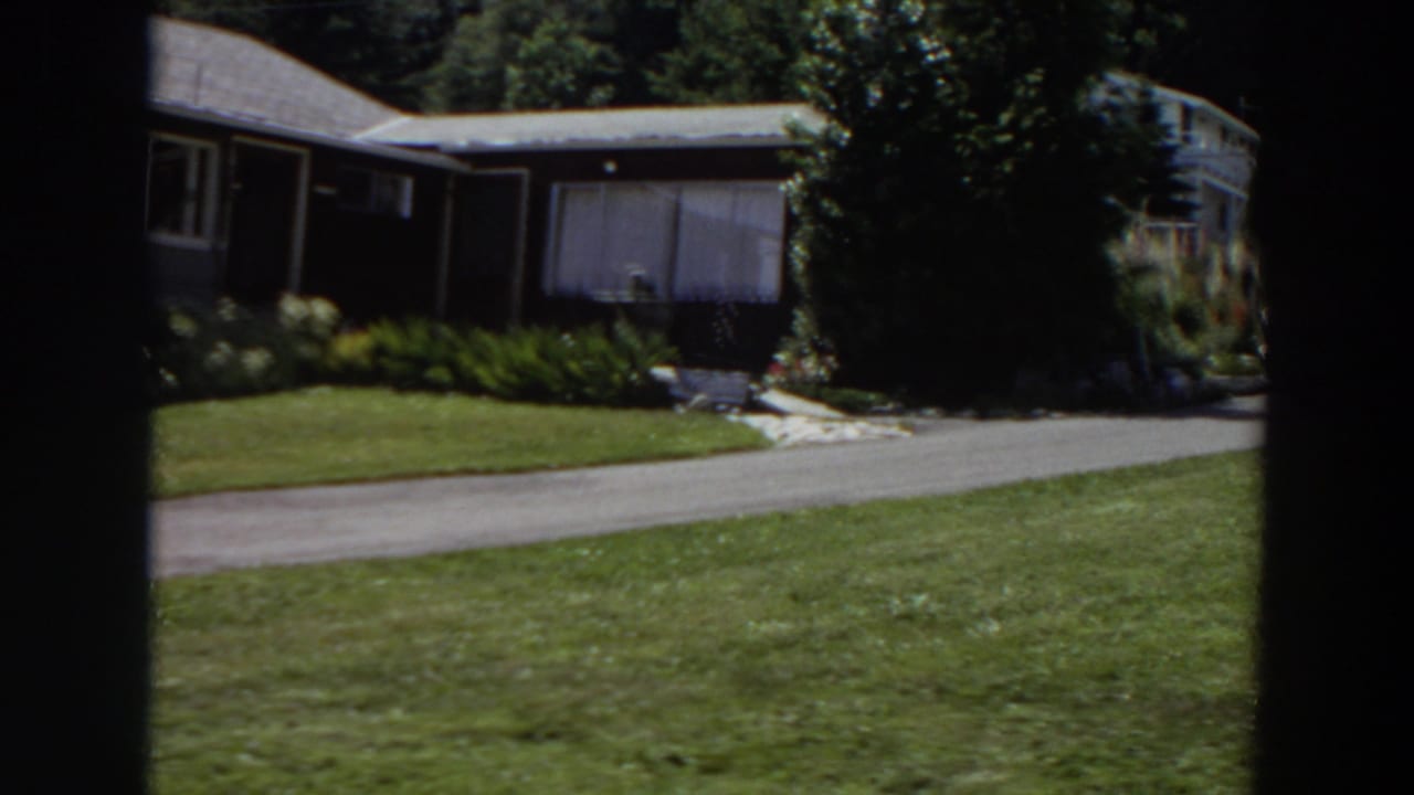 Dark-framed view of a low house, lawn, trees, and driveway before the car enters.