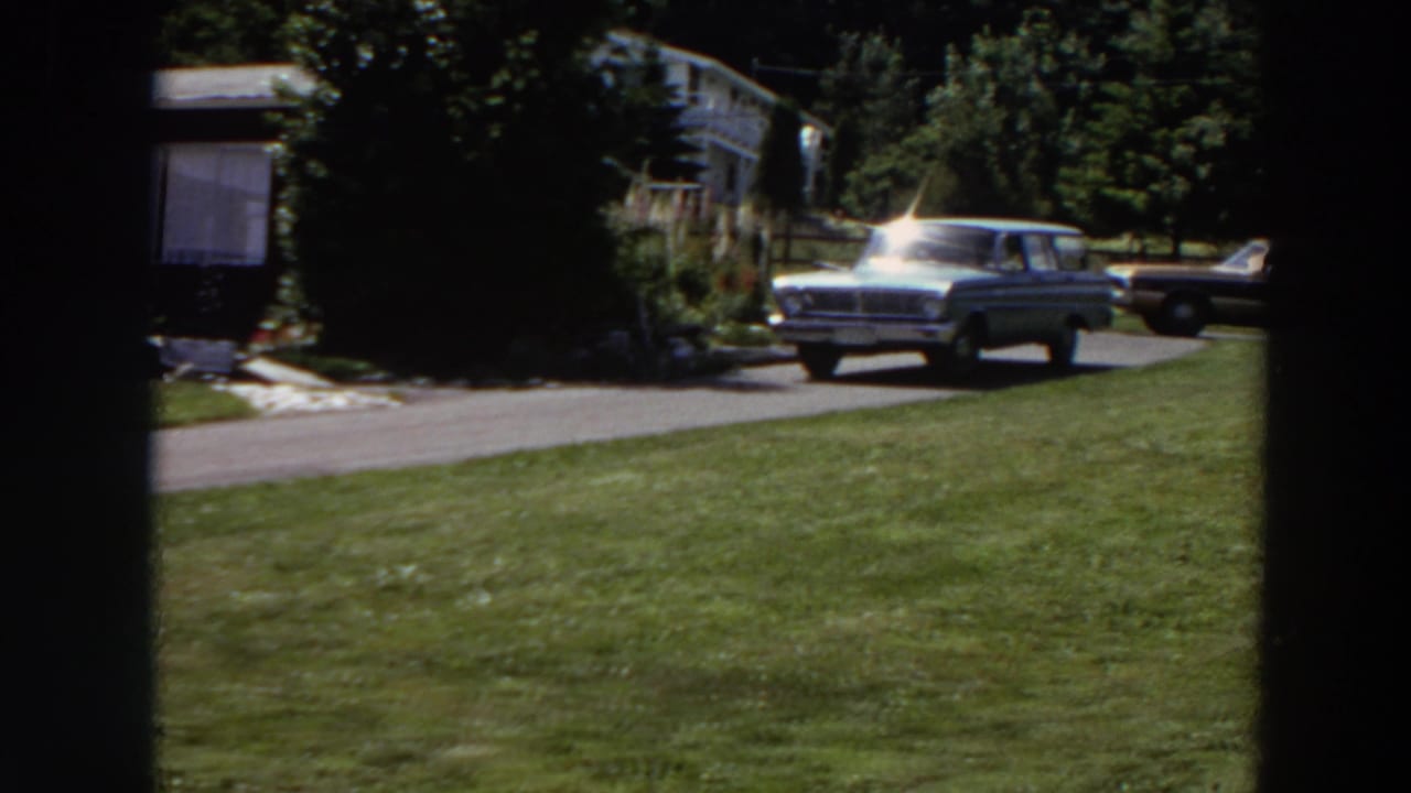 The light station wagon moves down the driveway past the house, with another vintage car parked behind it in the sunlit yard setting.