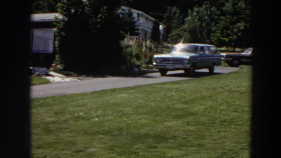 The light station wagon moves down the driveway past the house, with another vintage car parked behind it in the sunlit yard setting.