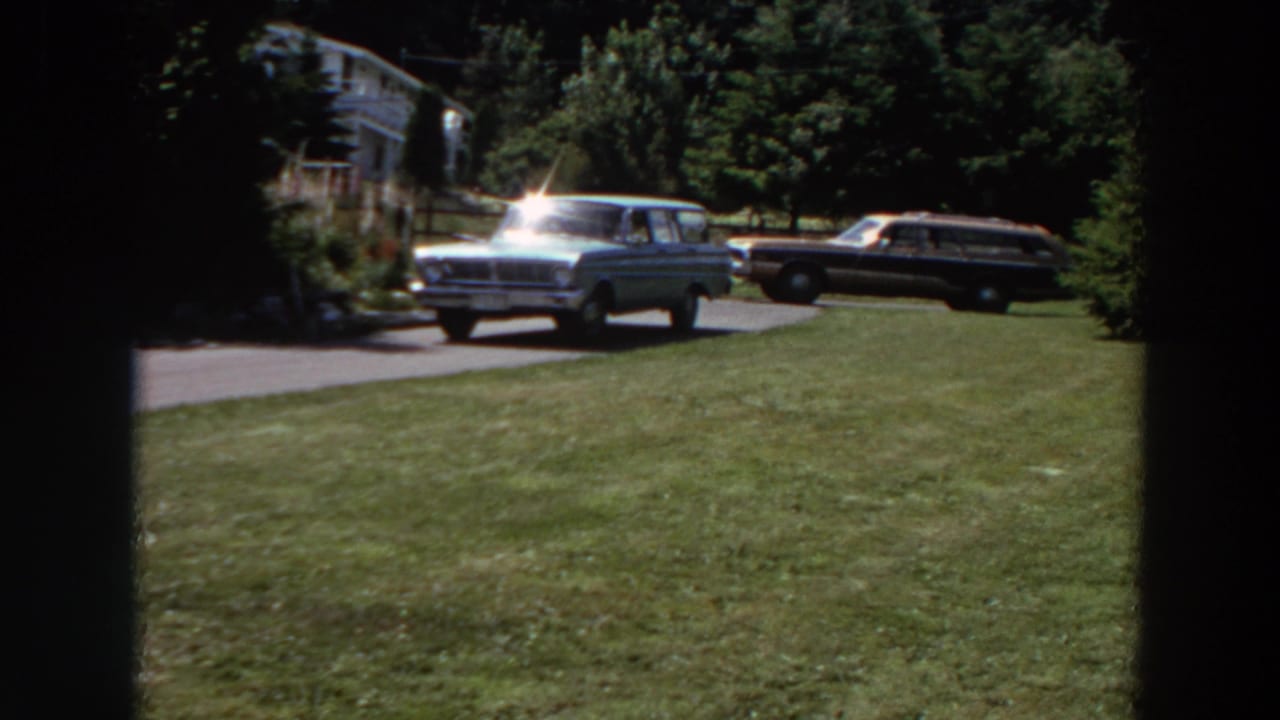 Near the end of the 1964 Washington clip, the station wagon continues along the residential driveway beside lawn, trees, and a parked car.
