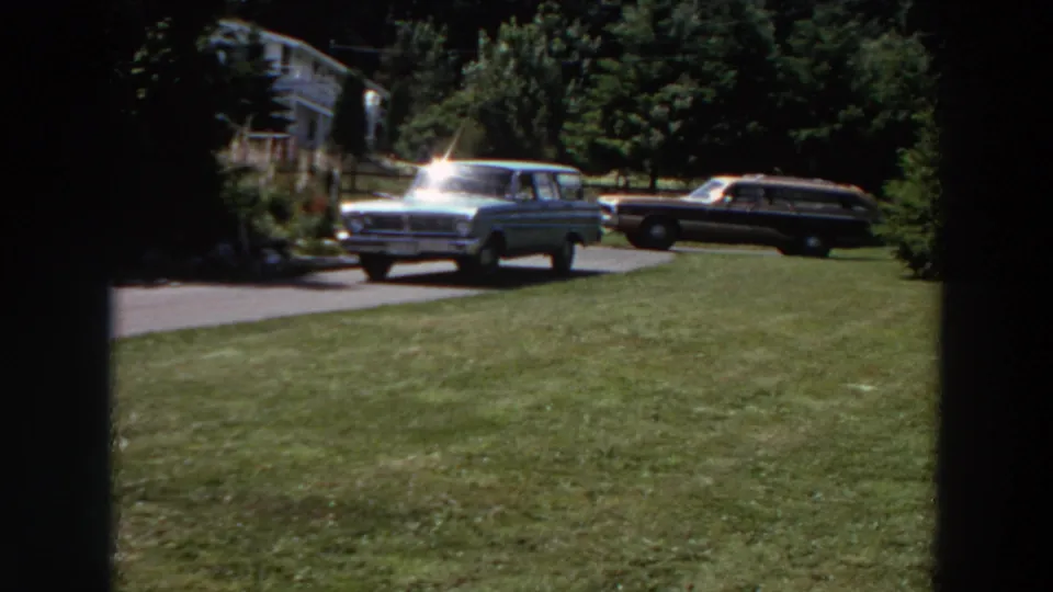 Near the end of the 1964 Washington clip, the station wagon continues along the residential driveway beside lawn, trees, and a parked car.