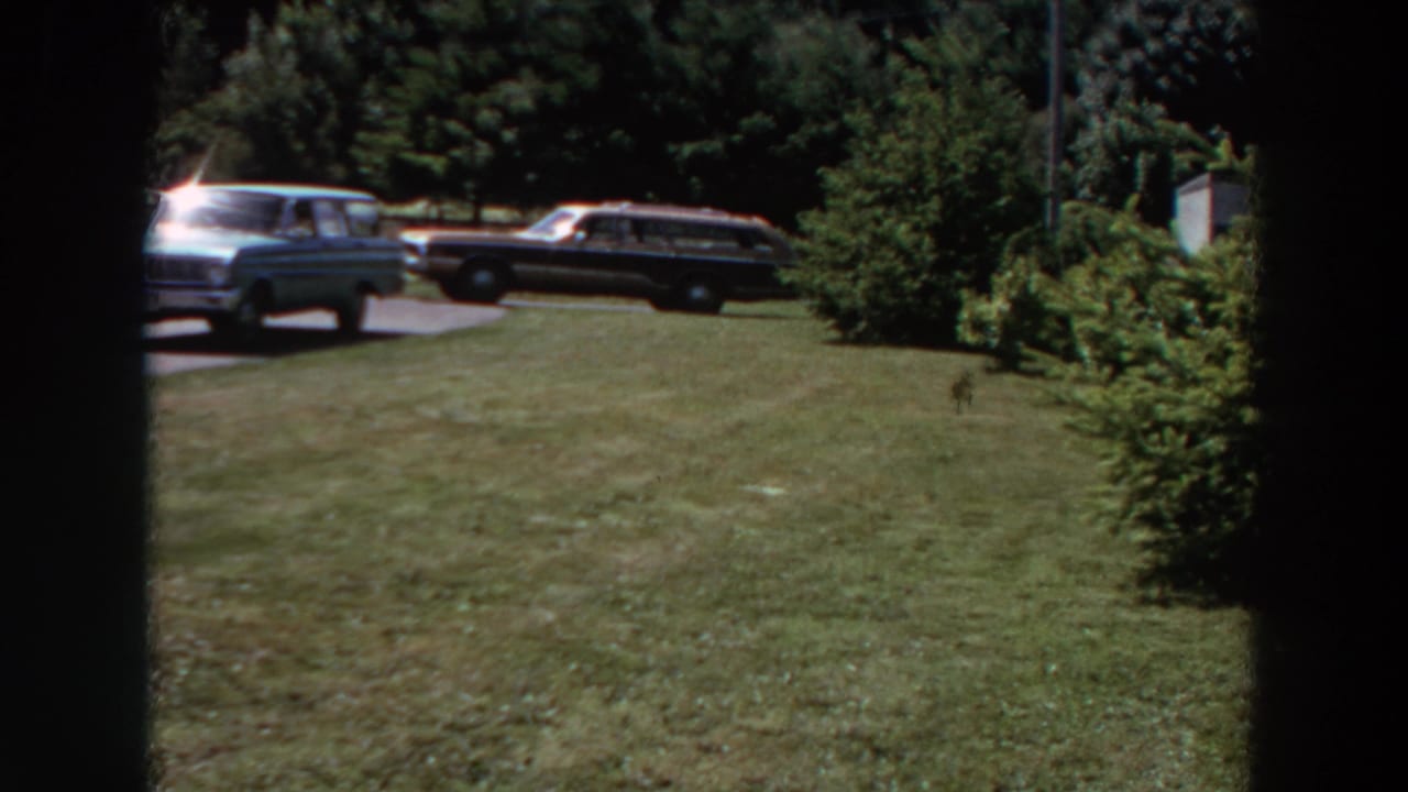 1964 Washington home-movie view of a broad suburban lawn, two parked station wagons, shrubs, trees, and partial residential structures.