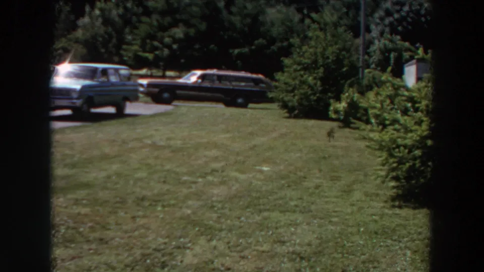1964 Washington home-movie view of a broad suburban lawn, two parked station wagons, shrubs, trees, and partial residential structures.