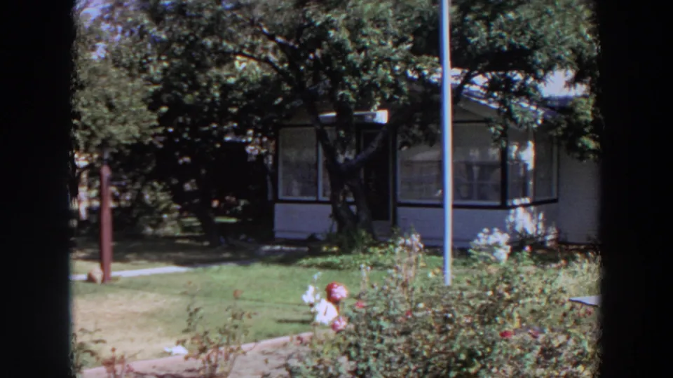 1964 Washington home-movie frame showing a white house front, enclosed porch, lawn, rose bushes, and shaded trees.