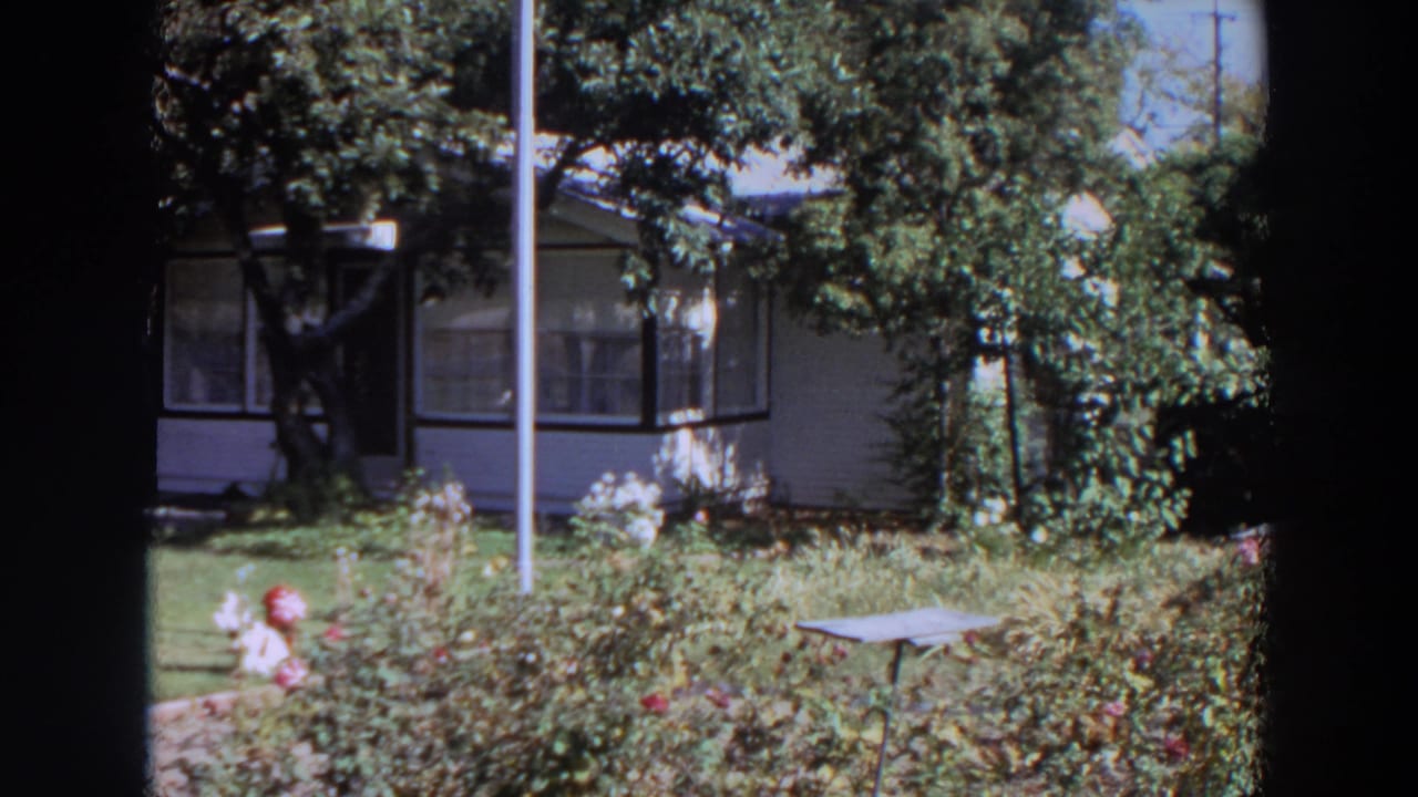 1964 Washington home-movie still of a shaded residential porch and front garden with lawn, shrubs, vertical pole, and small table.