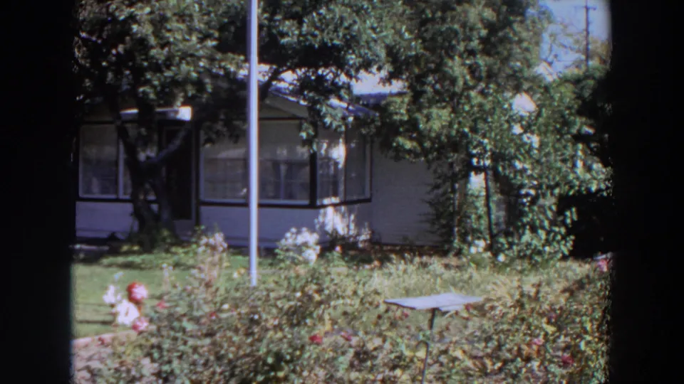 1964 Washington home-movie still of a shaded residential porch and front garden with lawn, shrubs, vertical pole, and small table.