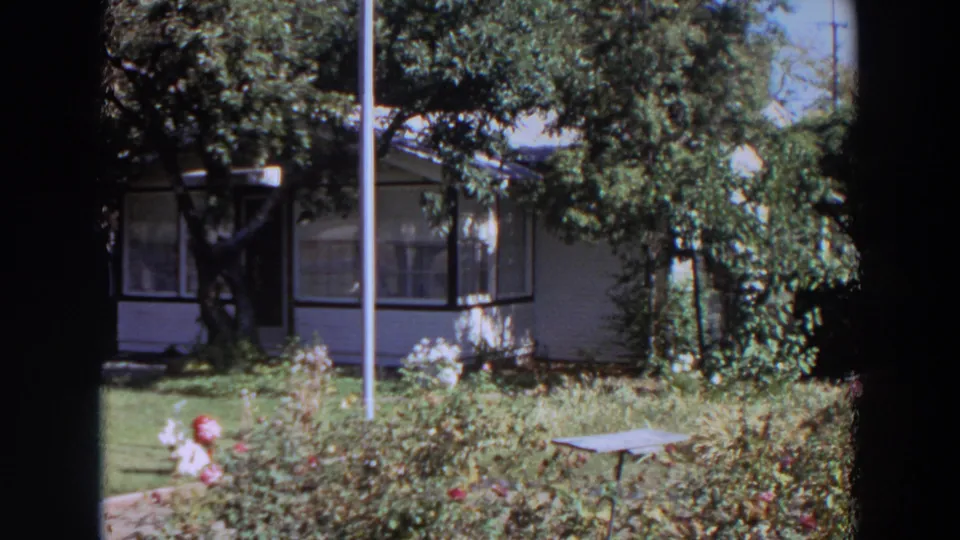 1964 Washington home-movie frame showing a white residential house, lawn, trees, and garden planting in bright daylight.
