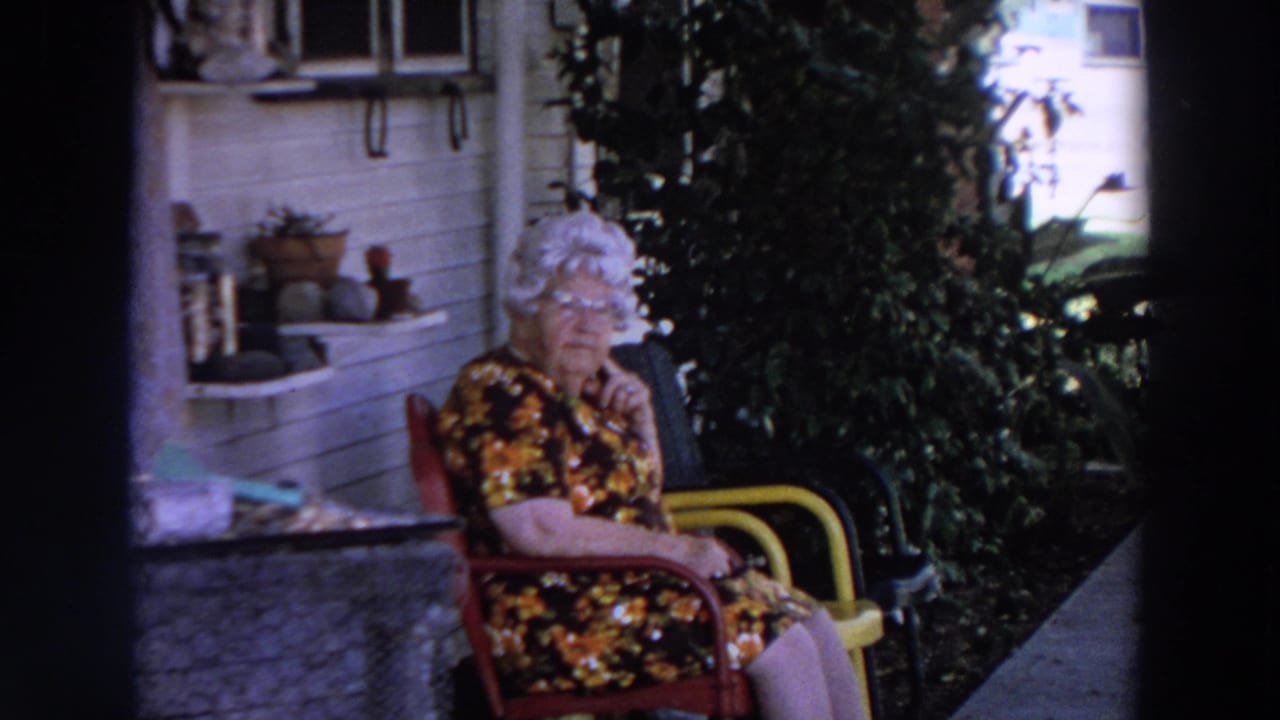 In Washington 1964 home-movie footage, an elderly woman in a floral dress sits outside near porch plants and white house siding.