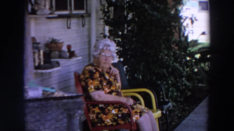 In Washington 1964 home-movie footage, an elderly woman in a floral dress sits outside near porch plants and white house siding.