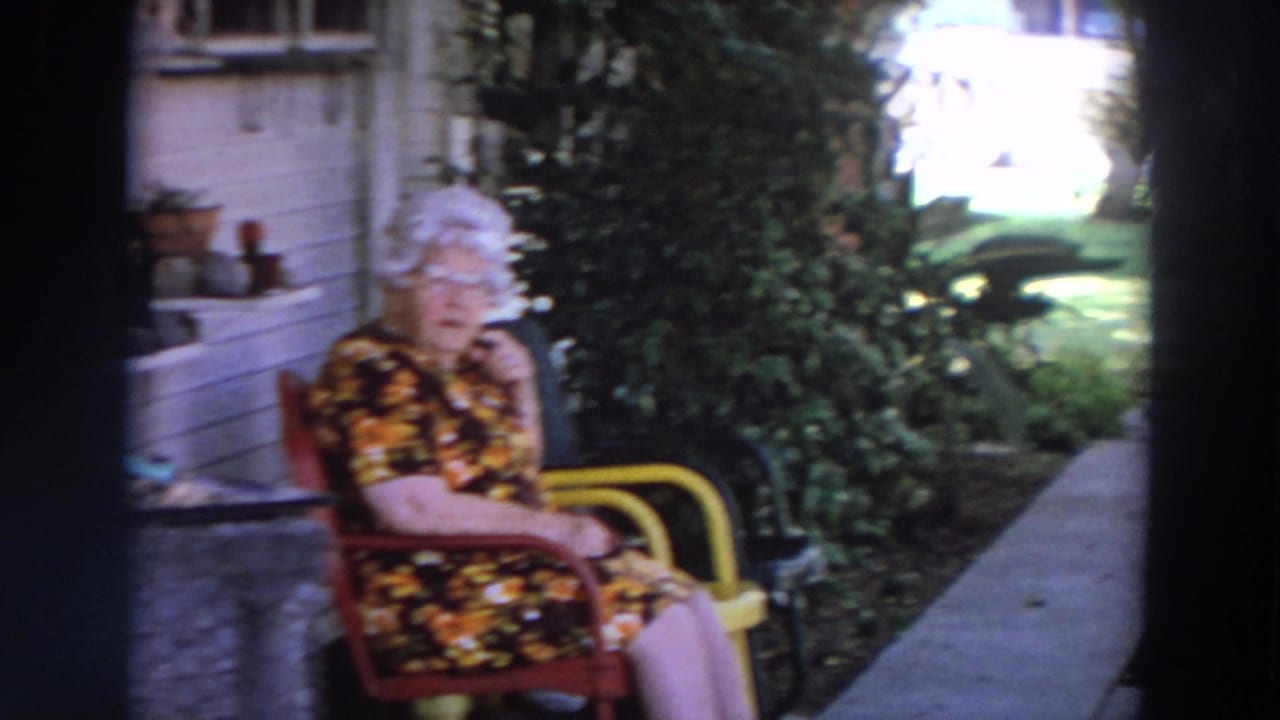 The seated woman, wearing glasses and a dark floral dress, is shown near a porch with greenery and a yellow chair arm visible.