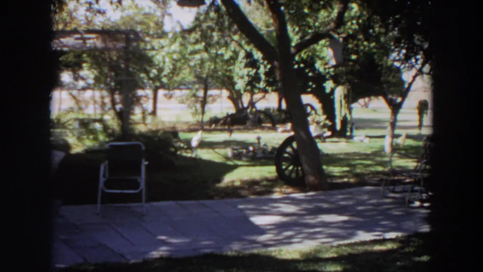 1964 Washington home-movie frame of a sunny backyard with patio, trees, wagon wheel, and an indistinct distant figure.