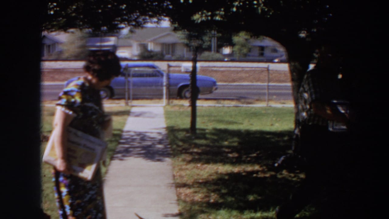 A woman in a patterned dress holds printed material near a sidewalk while a man carries a box by a street with a blue car.
