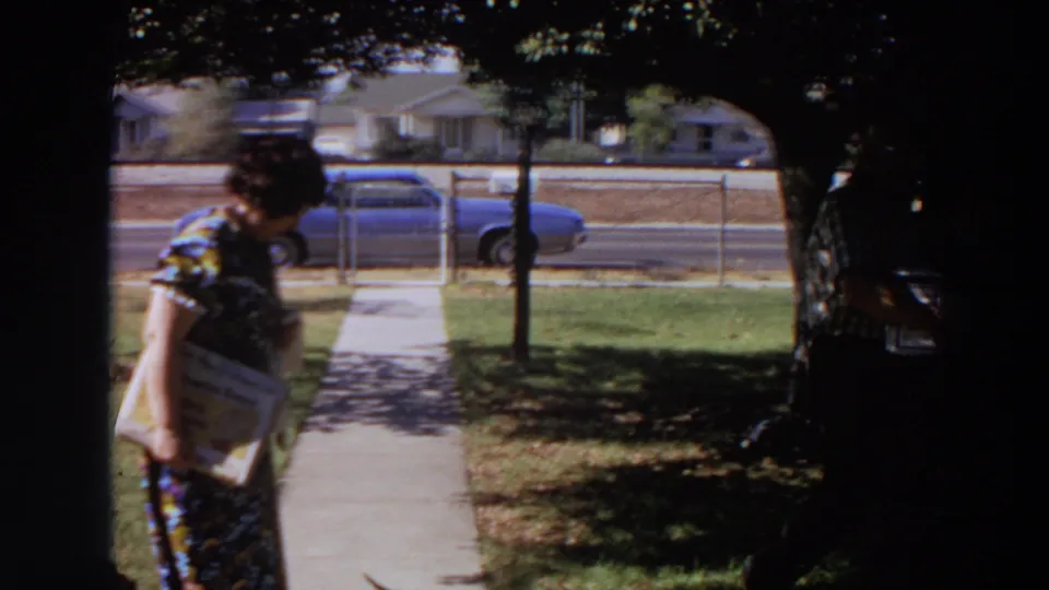 A woman in a patterned dress holds printed material near a sidewalk while a man carries a box by a street with a blue car.