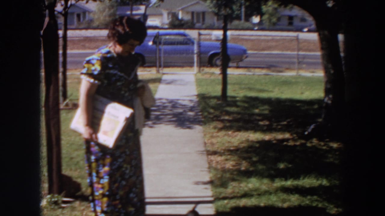 In 1964 Washington footage, a woman in a floral dress walks along a suburban sidewalk with a book or papers in hand.