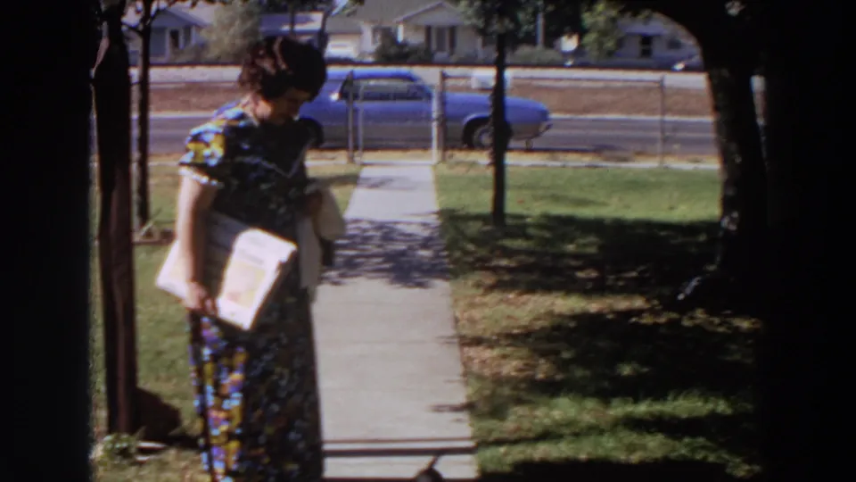 In 1964 Washington footage, a woman in a floral dress walks along a suburban sidewalk with a book or papers in hand.