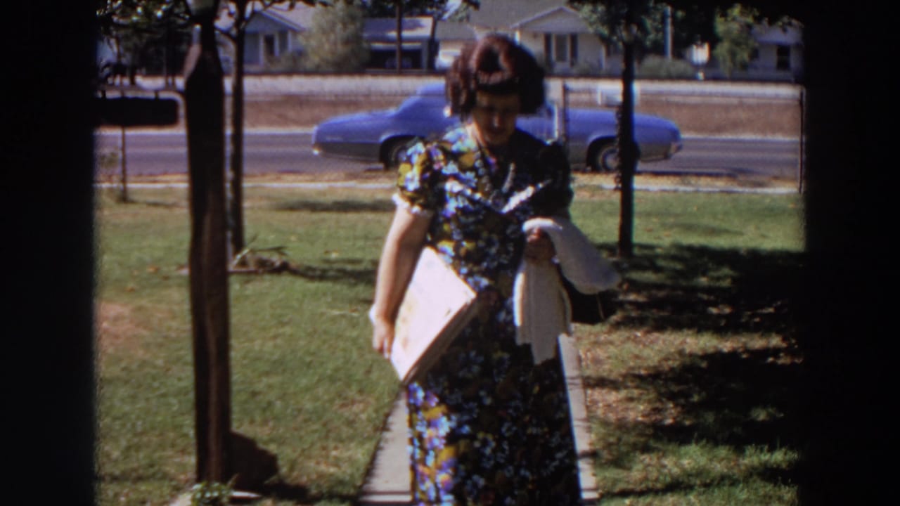The woman approaches the camera in a 1964 Washington suburban yard scene, with a blue car and chain-link fence behind her.