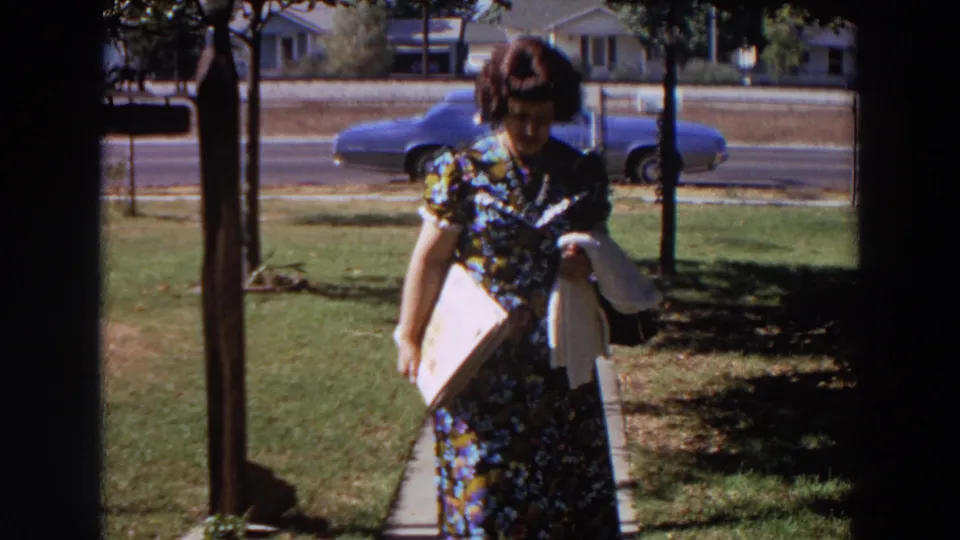 The woman approaches the camera in a 1964 Washington suburban yard scene, with a blue car and chain-link fence behind her.