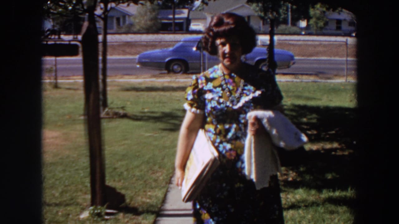 A woman in mid-1960s dress walks toward the camera in a Washington residential setting, with lawn, sidewalk, fence, and parked car visible.