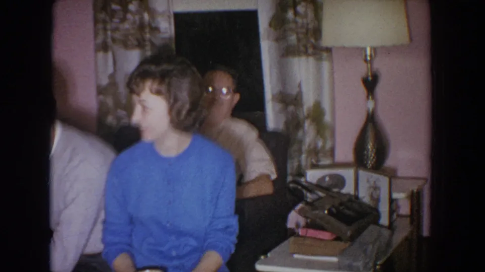 1965 Fallston, Maryland home interior with a woman in a blue cardigan seated near a lamp, curtains, and side table objects.