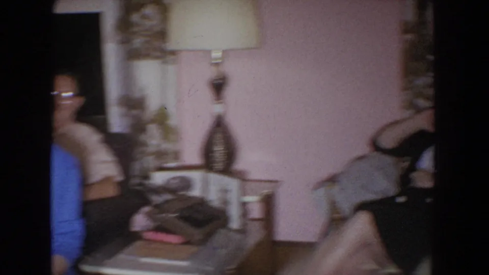 1965 Fallston, Maryland home-movie frame with motion blur across a pink living room, lamp, side table, and partially seen adults.