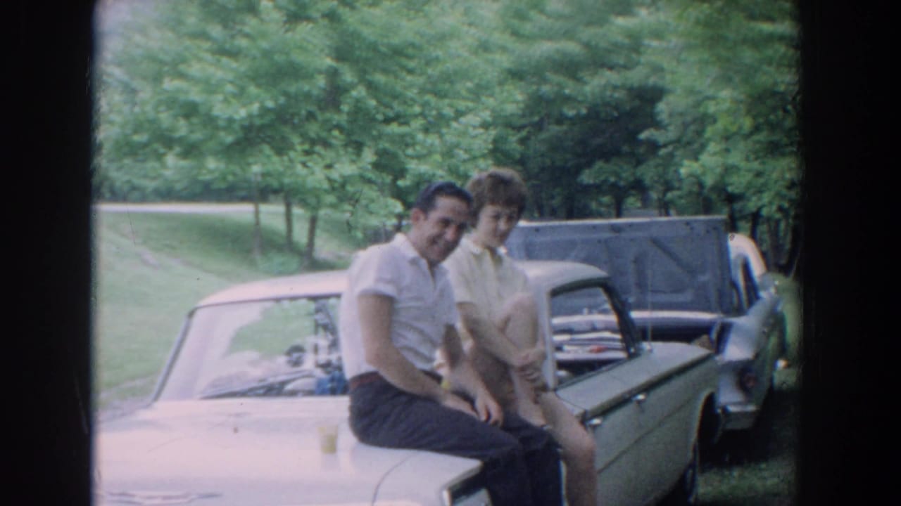 Smiling couple seated on a light-colored car with another car behind them.
