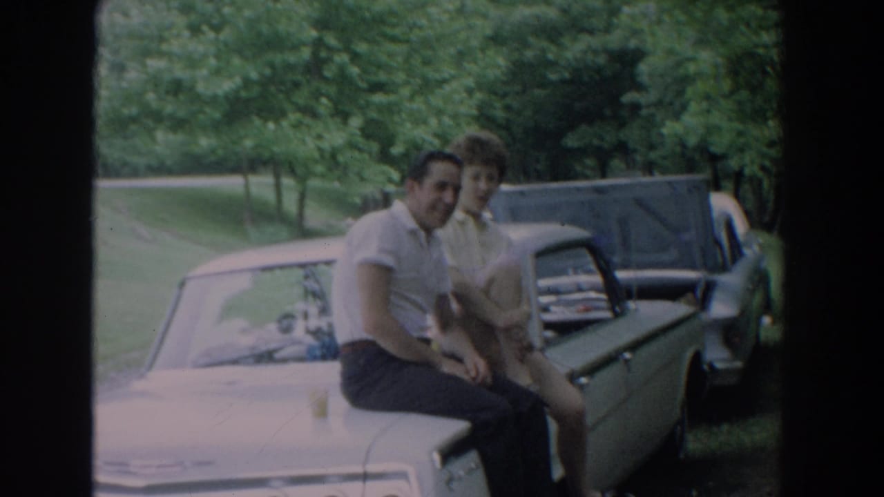 Couple seated together on a car hood with trees and an open car hood behind them.