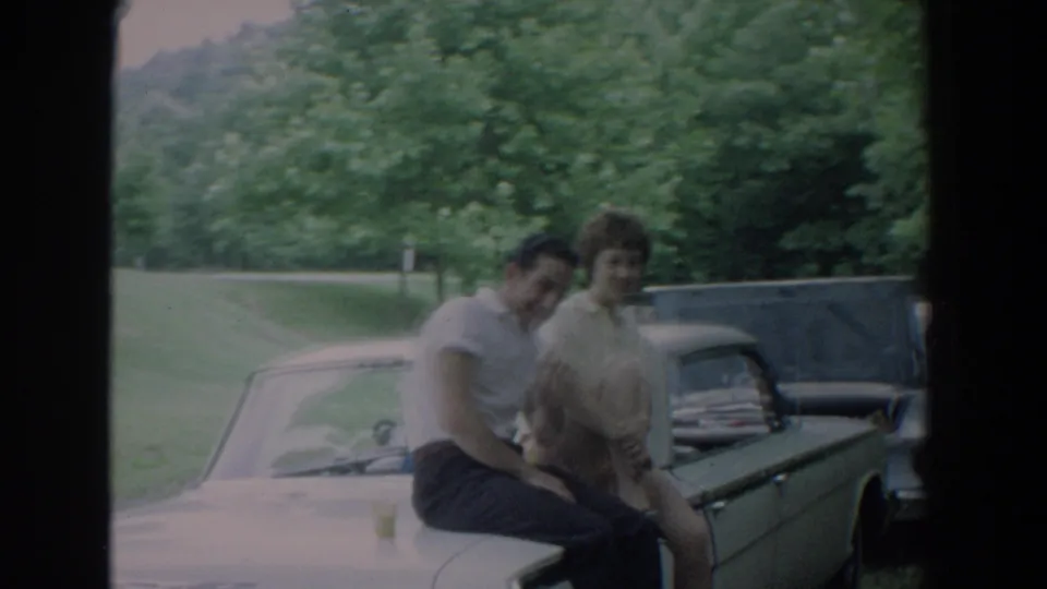 A soft 8mm frame shows the Fallston couple moving slightly while seated on the car hood in a green roadside setting.