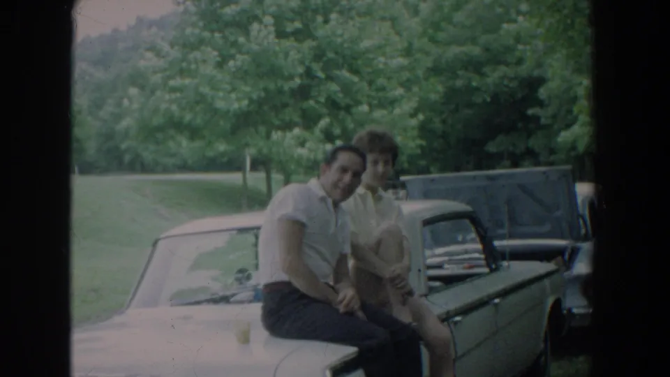 The couple looks toward the camera from the hood of a light-colored vintage car in the 1965 Fallston home-movie clip.