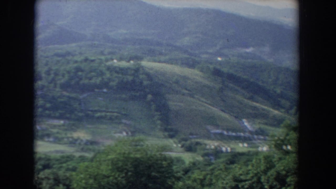 Soft distant view of green hills, pale fields, trees, and a few small homes.