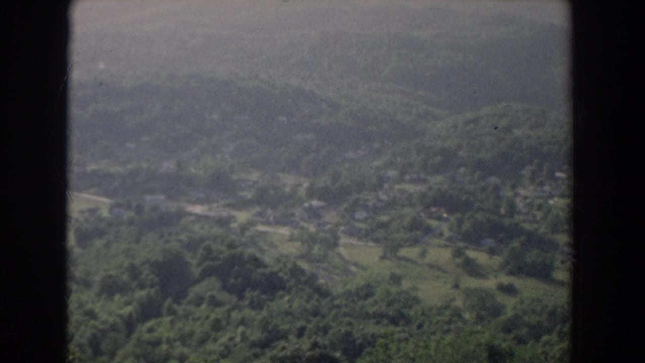 Aerial landscape with wooded slopes, fields, roads, and small buildings below.