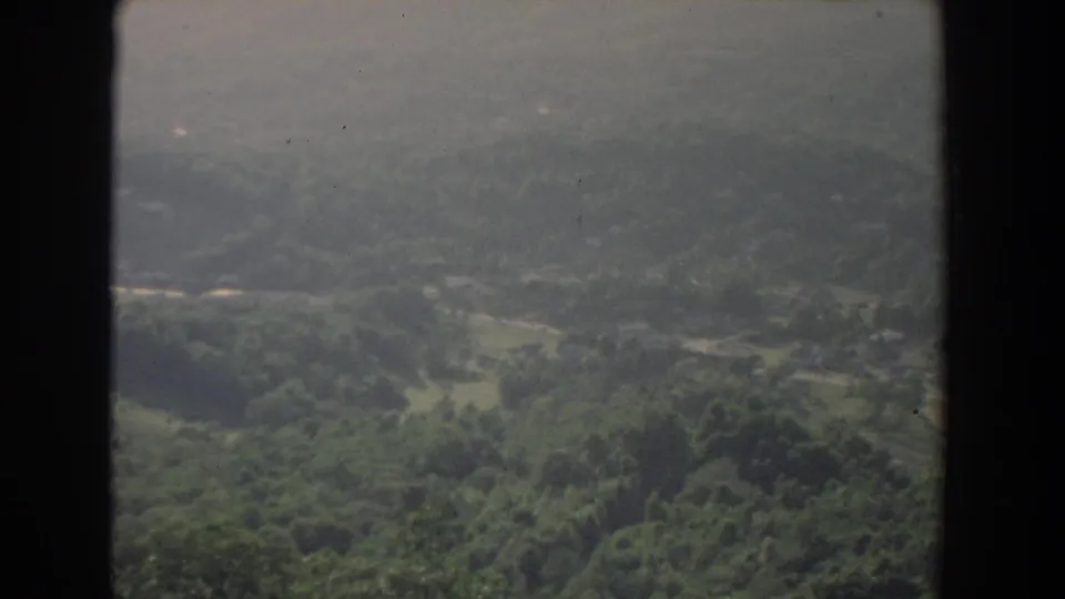 1965 8mm view over the Fallston area, with tree-covered slopes, open fields, faint roads, and soft film grain.