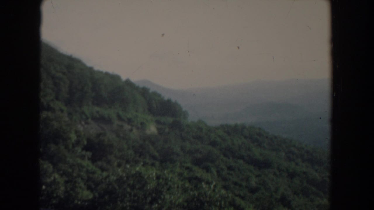 Forested hillside and distant hazy ridges seen through a dark vertical obstruction.