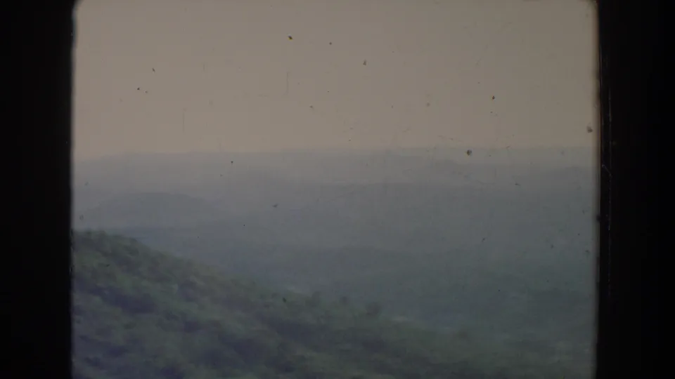 A hazy rural valley and low ridges fill the frame, with pale sky and visible 8mm film borders from the 1965 home-movie clip.