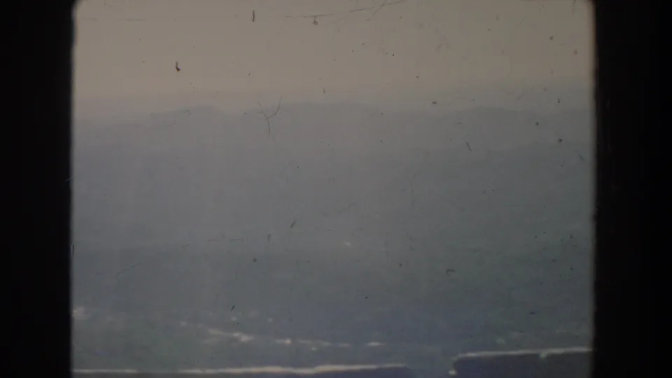 1965 home-movie frame with a fog-softened valley or water surface, faint shoreline forms, pale sky, and visible film scratches.