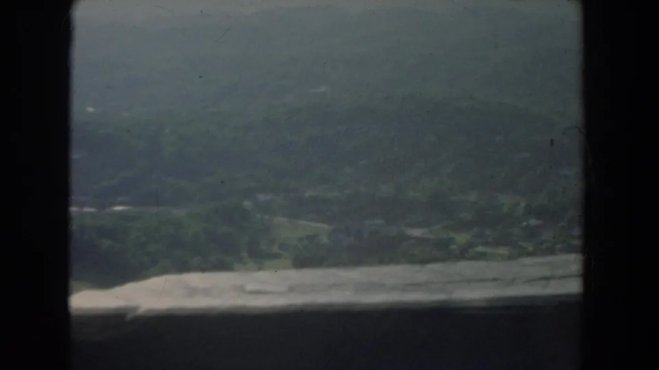 Hazy 1965 8mm view of wooded hills and a green valley, partially blocked by a dark foreground edge and film-frame border.