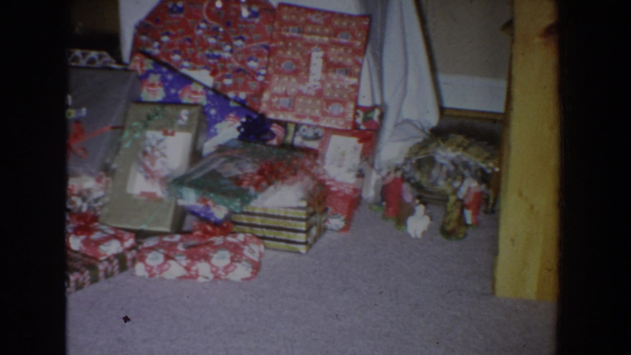 Wrapped Christmas presents stacked on a carpeted floor beside small holiday figurines.